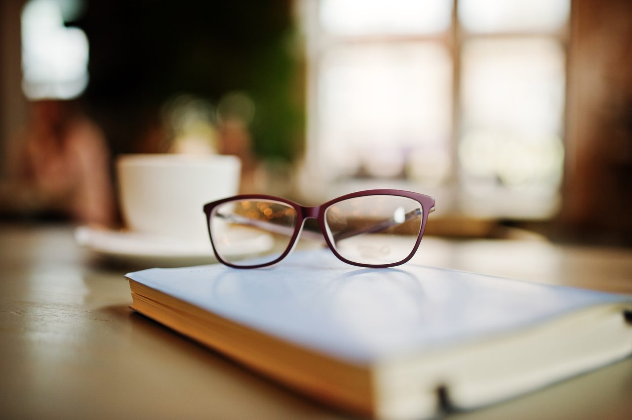 Rote Brille auf einem Buch, dahinter eine Kaffeetasse