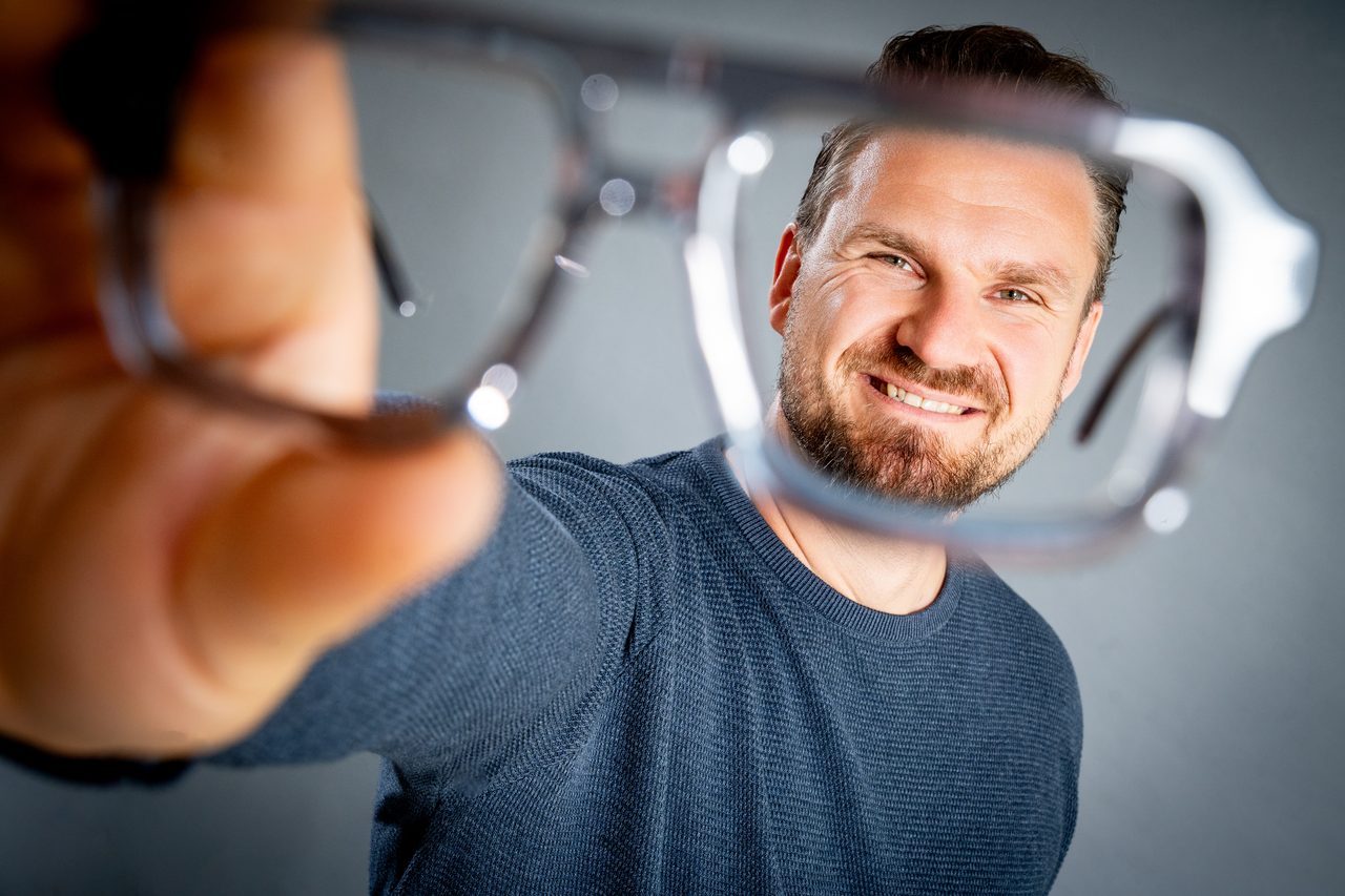 Marian Ried hält lachend eine Brillenfassung vor die Kamera und schaut hindurch