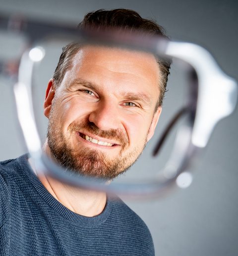 Marian Ried hält lachend eine Brillenfassung vor die Kamera und schaut hindurch